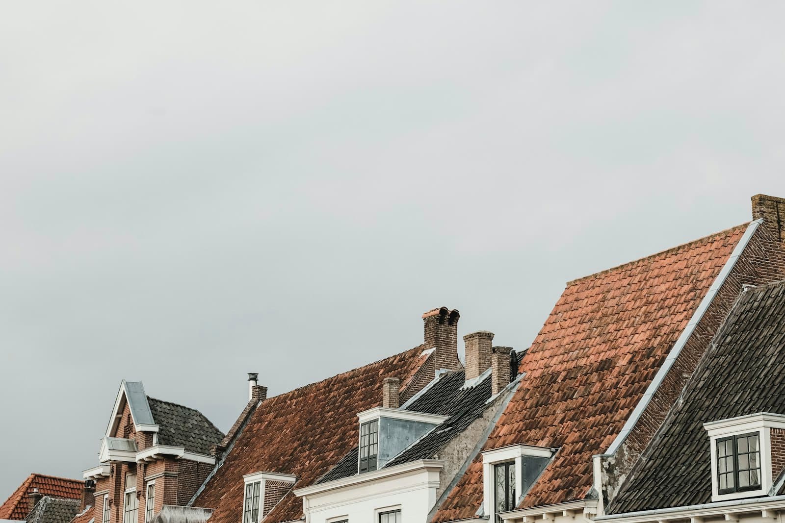 Nederlandse daken naast elkaar met verweerde rode pannen, donkere pannen, dakkapellen en schoorstenen onder een bewolkte lucht